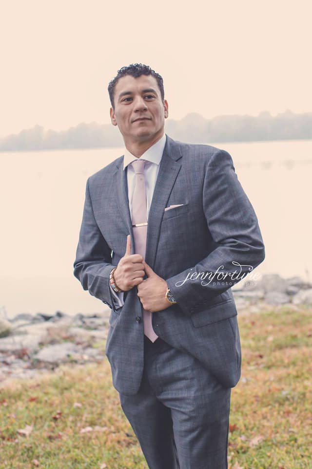 A man in a gray suit and pink tie stands by a body of water, adjusting his suit jacket with a confident expression.